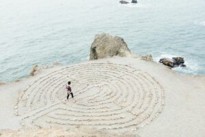 A person walks through a circular stone labyrinth on a coastal cliff, reflecting on relationships, with the ocean and rocky formations in the background under an overcast sky.