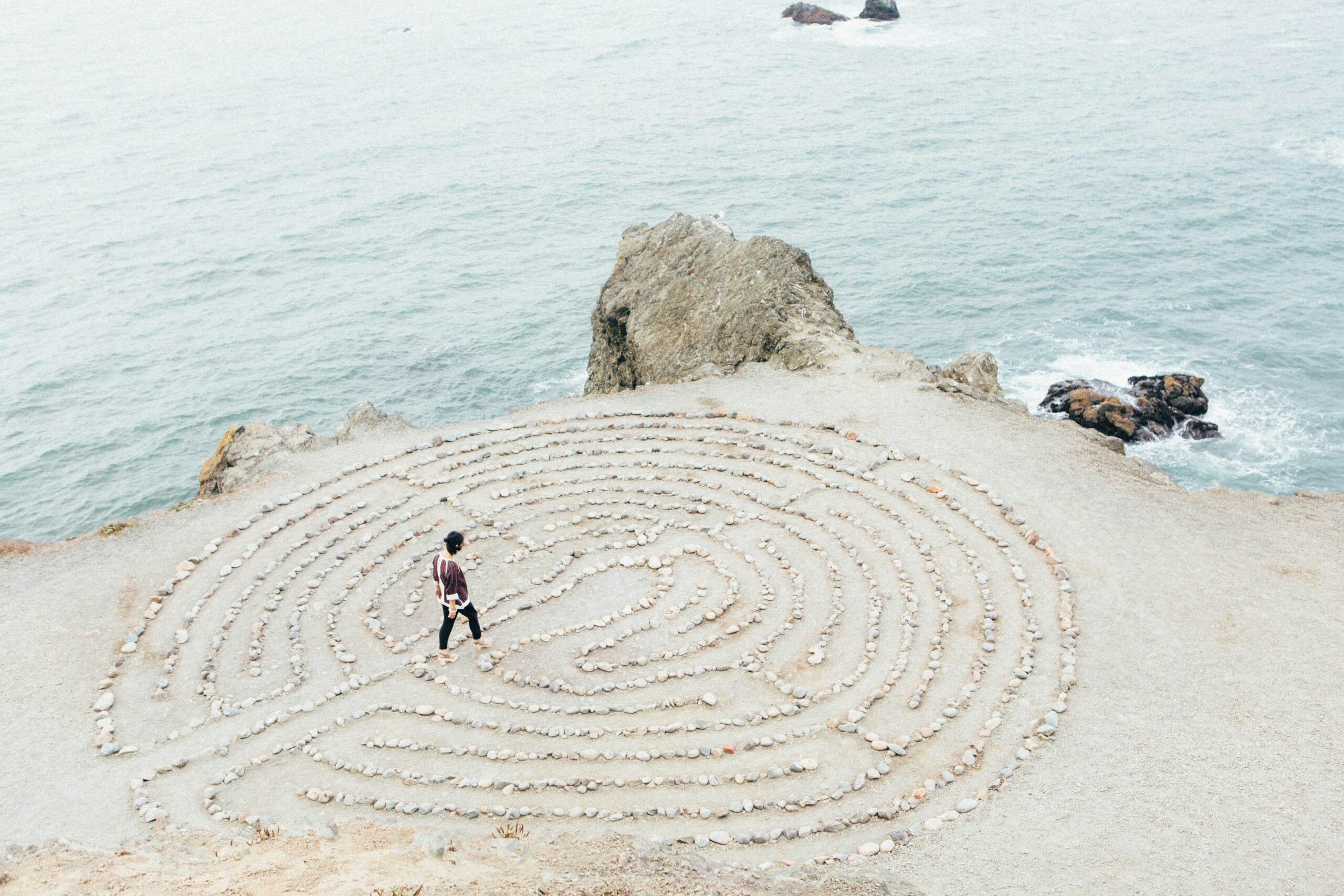 A person walks through a circular stone labyrinth on a coastal cliff, reflecting on relationships, with the ocean and rocky formations in the background under an overcast sky.