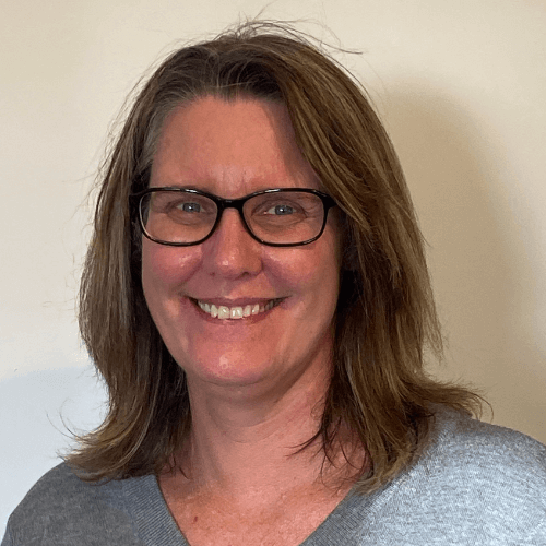 A woman with shoulder-length brown hair and glasses smiles at the camera. She is wearing a gray top and standing in front of a plain, light-colored background, reflecting the welcoming approach found at About Khiron Clinics.