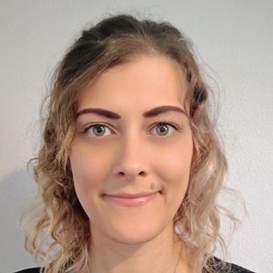 A woman with curly, light brown hair and green eyes smiles slightly at the camera. She is standing in front of a plain white wall, wearing a black top—an authentic portrait echoing the welcoming spirit found at Khiron Clinics.