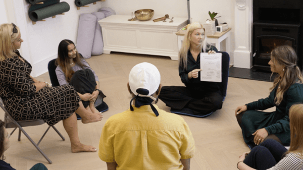 A group of adults sits in a cozy room, gathered in a circle. One woman holds up a piece of paper and discusses What is Trauma, while others listen attentively. Yoga mats and relaxation items are visible in the background.