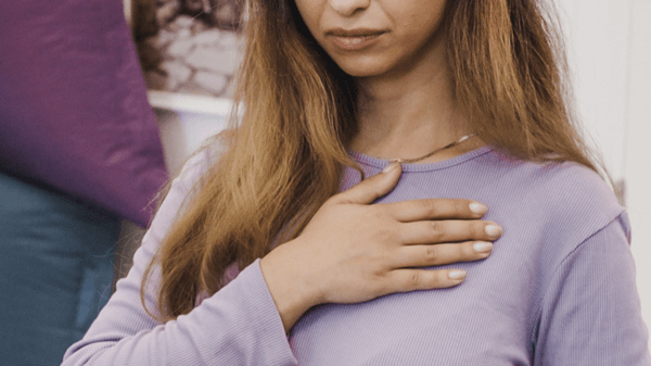 A woman wearing a lavender long-sleeve shirt places her right hand over her chest, appearing thoughtful or contemplative—perhaps reflecting on the question, "What is Trauma?.