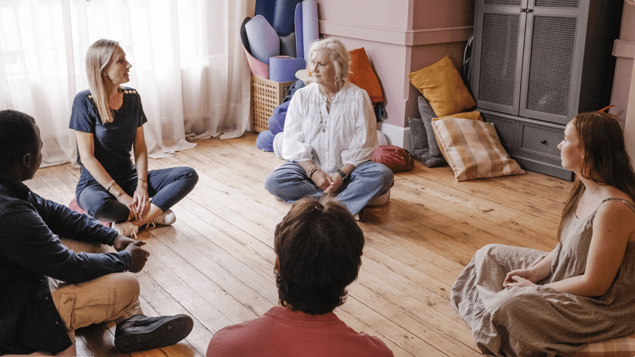 Five people sit in a circle on a wooden floor in a cosy, sunlit room with cushions and yoga mats, engaging in a group conversation about What is Trauma and supporting each other through open discussion.