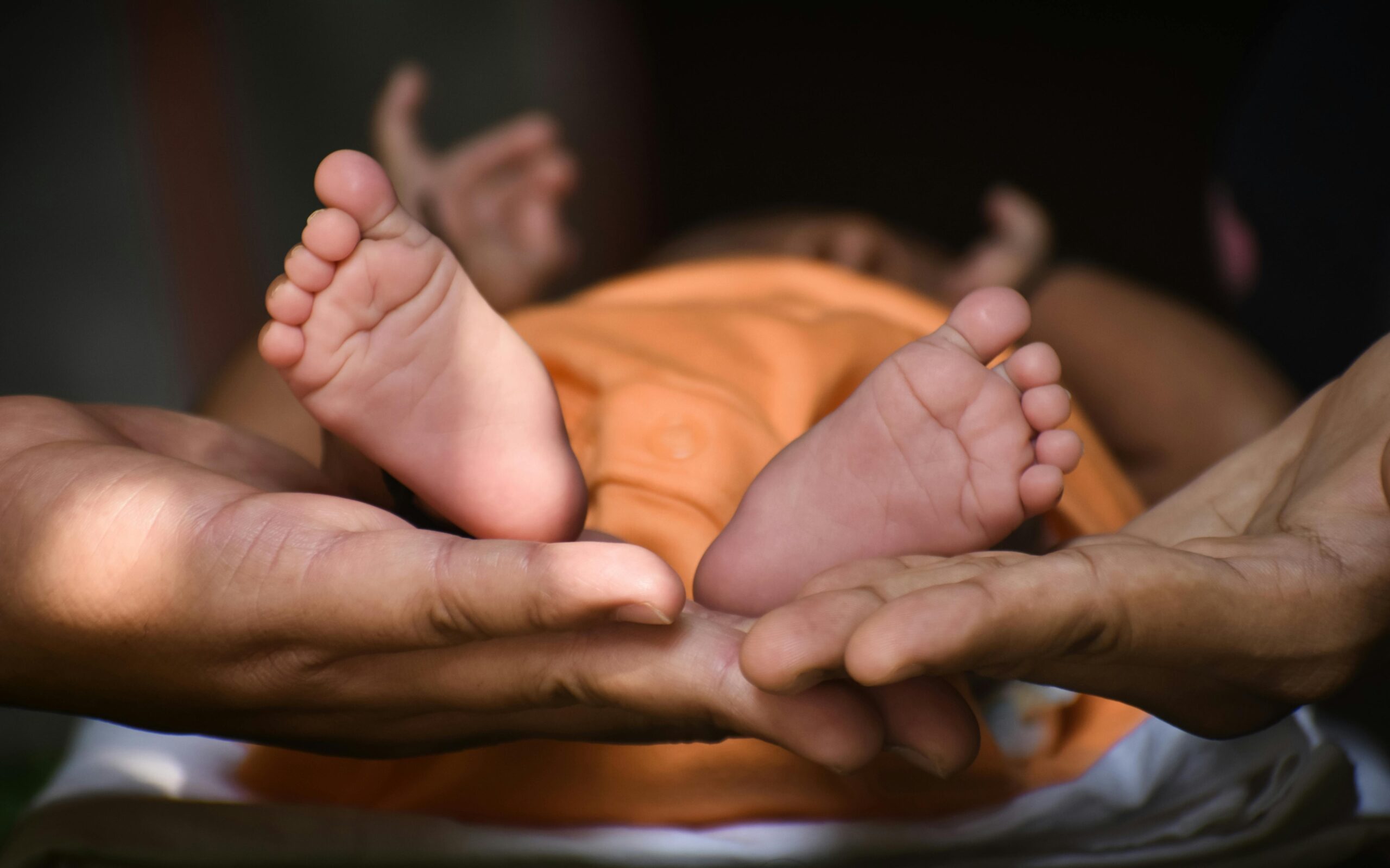 Close-up of adult hands gently cradling the tiny feet of a baby lying down in an orange outfit, with the baby’s legs and toes in focus against a dark, blurred background—capturing a tender moment that can help heal postnatal depression.