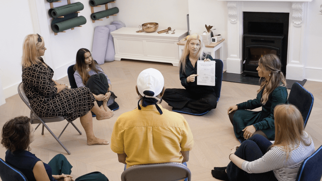 A group of people sit in a circle in a bright room during a Trauma Workshop London, some on chairs and others on the floor, as a woman holds up a piece of paper and leads the discussion. Yoga mats and cushions are stacked in the background.