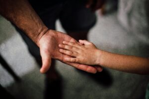 A child’s hand rests gently on an adult’s open palm, symbolising care, connection, and trust—a quiet moment that hints at healing from generational trauma. The softly blurred background keeps the focus on the hands.