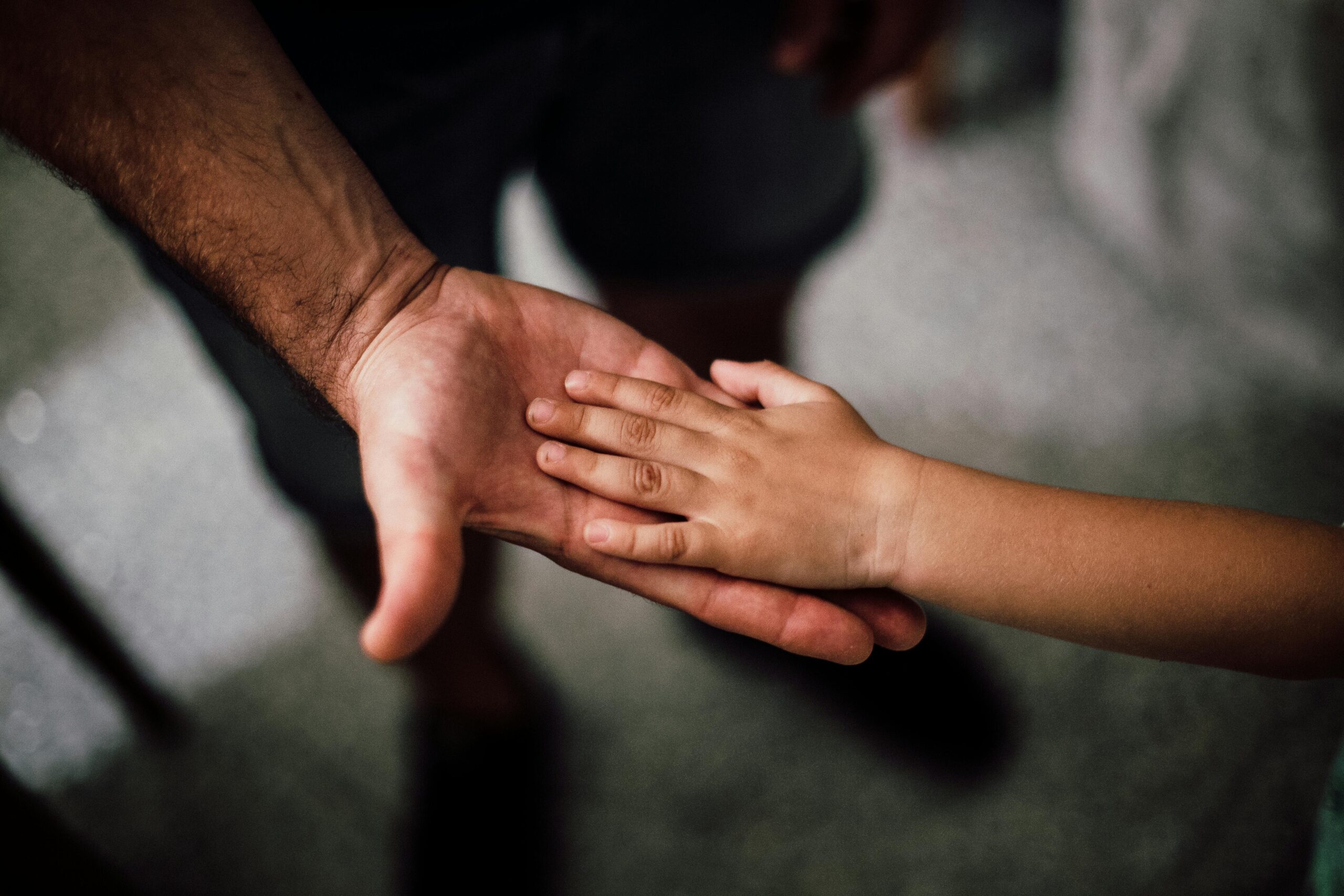 A child’s hand rests gently on an adult’s open palm, symbolising care, connection, and trust—a quiet moment that hints at healing from generational trauma. The softly blurred background keeps the focus on the hands.