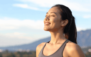 A woman with long dark hair in a ponytail stands outdoors, smiling with her eyes closed, enjoying the sunlight, a moment of healing as she embraces peace and calm beneath the blue sky. She wears a grey vest top, with a blurred landscape behind her.