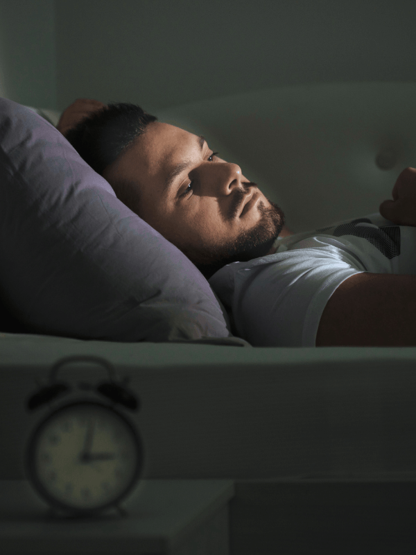 A man lies awake in bed at night, resting his head on a pillow and staring thoughtfully at the ceiling. An analogue alarm clock on a bedside table shows the time as nearly 3:00. The room is dimly lit.
