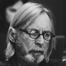 Black and white photo of an older man with fair hair and a beard, wearing round spectacles and a dark jumper, looking down with a focused expression—perfect for illustrating the compassionate care at Khiron Clinics.