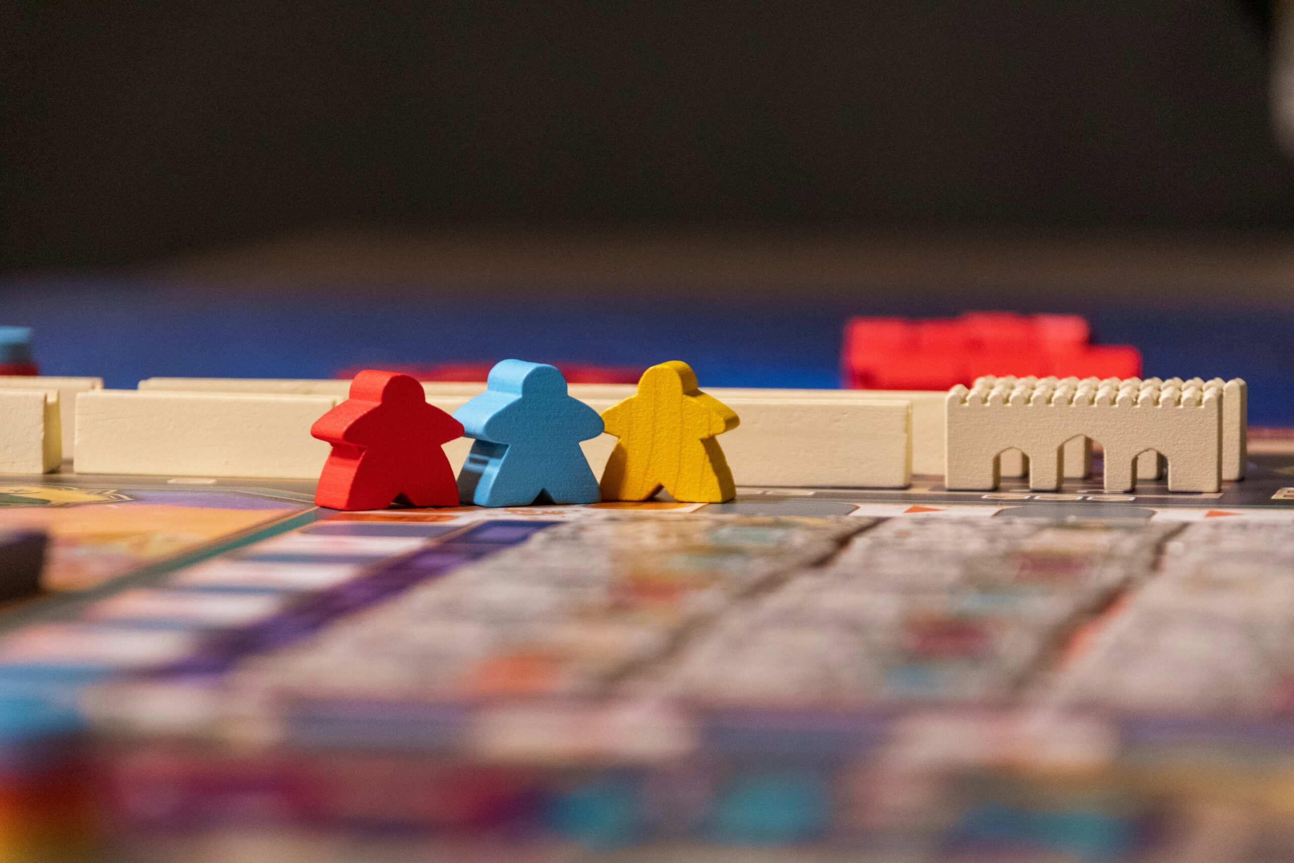 Three wooden game pieces—red, blue, and yellow—stand side by side on a colourful board near a white plastic wall and arch, symbolising the anxiety and tension often felt during challenging board games. The background is blurred.