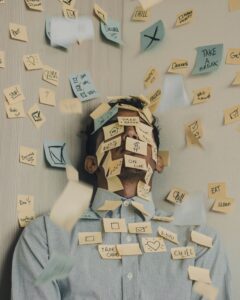 A person leans against a wall, their face and body covered in colourful sticky notes with doodles and words. More notes fall around them, capturing the chaos of stress and distress through a whirlwind of frantic reminders.