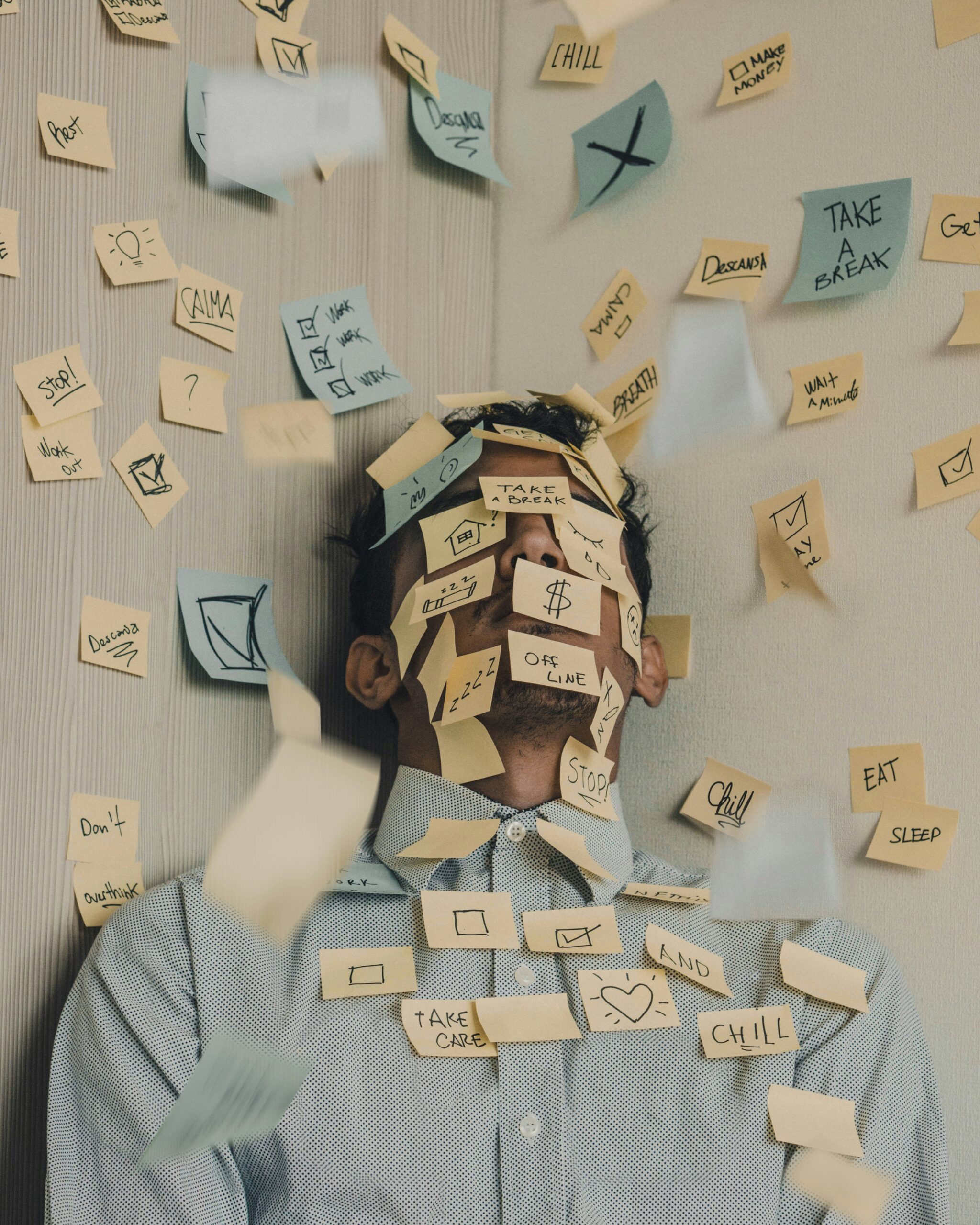 A person leans against a wall, their face and body covered in colourful sticky notes with doodles and words. More notes fall around them, capturing the chaos of stress and distress through a whirlwind of frantic reminders.