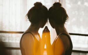 A woman with her hair in a bun stands close to a window, touching her forehead and hand to her reflection in the glass, as warm sunlight creates a soft, glowing effect—capturing a quiet moment of mental health reflection.
