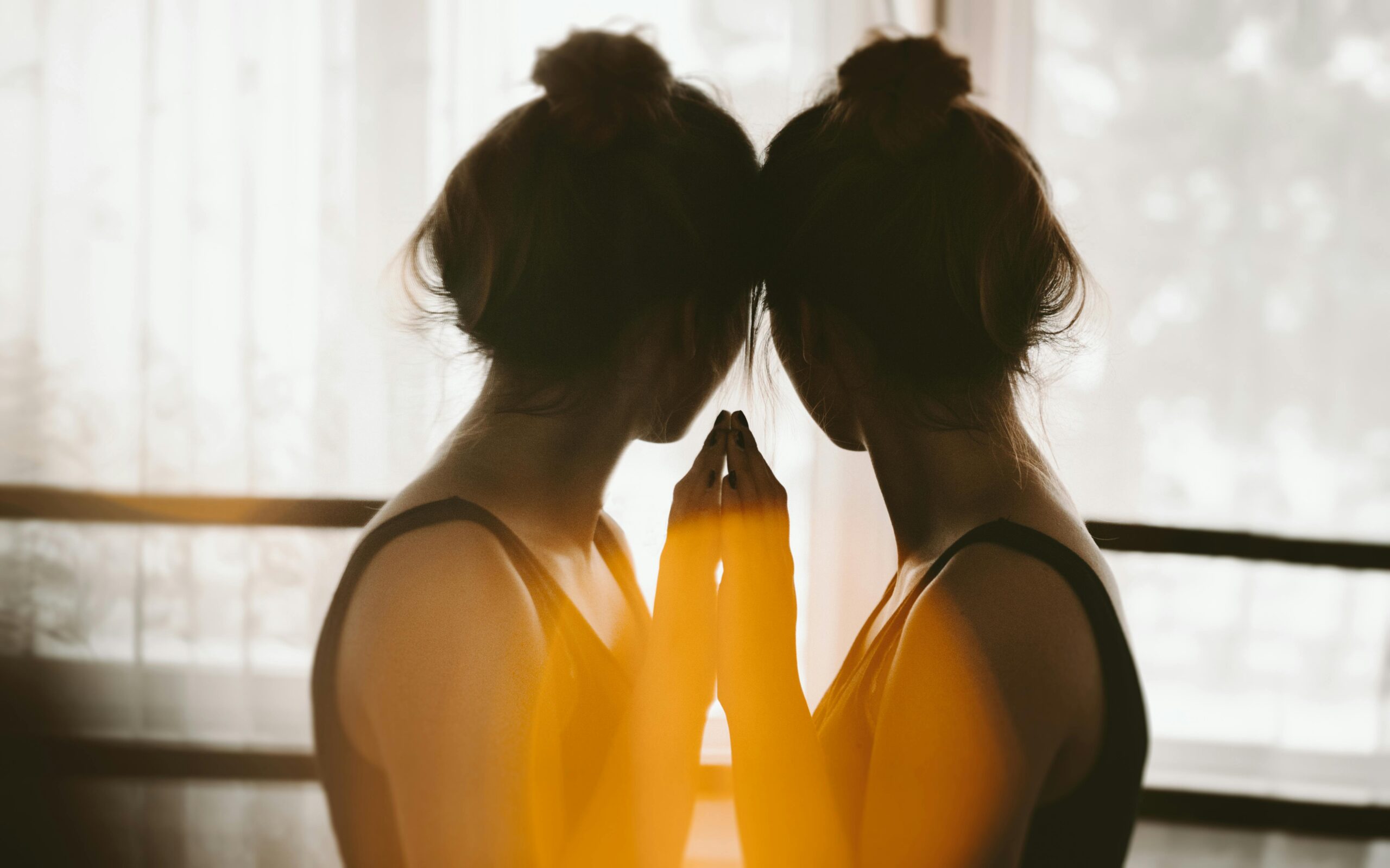 A woman with her hair in a bun stands close to a window, touching her forehead and hand to her reflection in the glass, as warm sunlight creates a soft, glowing effect—capturing a quiet moment of mental health reflection.