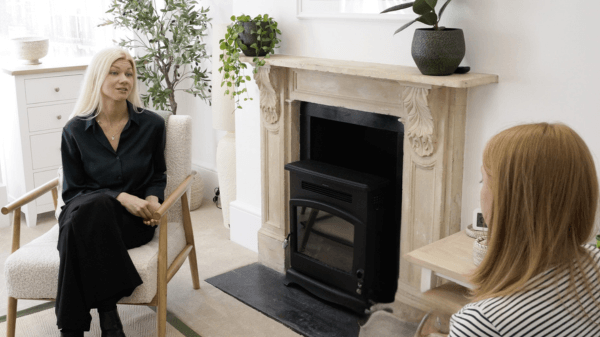 Two women sit facing each other, discussing what is trauma in a bright room with plants, a white fireplace, and neutral decor. One gestures as she speaks whilst the other listens attentively.