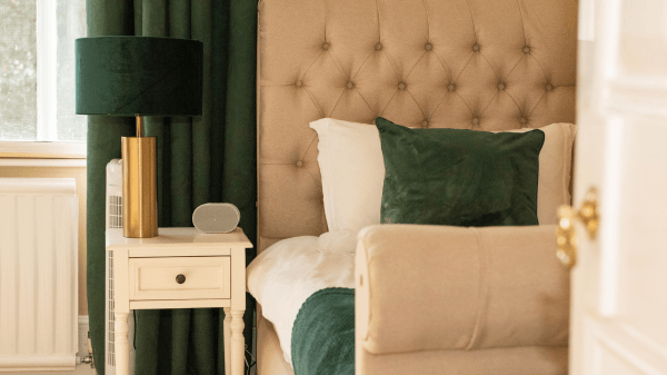 A cosy bedroom features a beige tufted headboard, white bedding with a green throw and pillow, a white bedside table with a modern black and gold lamp, and dark green curtains—creating a soothing retreat for anyone wondering what is trauma recovery.