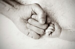 A close-up of an adult hand and a baby’s hand, both forming fists and gently touching each other, symbolising parenting and connection. The soft, light background adds warmth and tenderness to this heartfelt moment. It highlights that parenting can awaken old wounds.