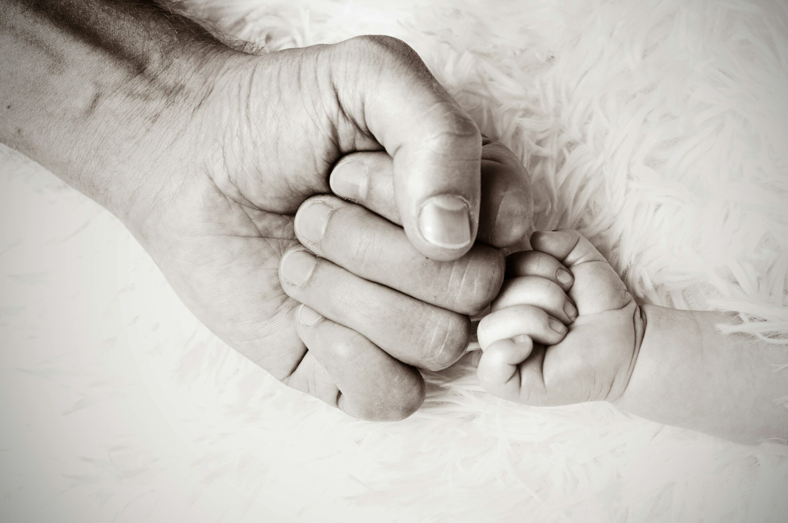 A close-up of an adult hand and a baby’s hand, both forming fists and gently touching each other, symbolising parenting and connection. The soft, light background adds warmth and tenderness to this heartfelt moment. It highlights that parenting can awaken old wounds.