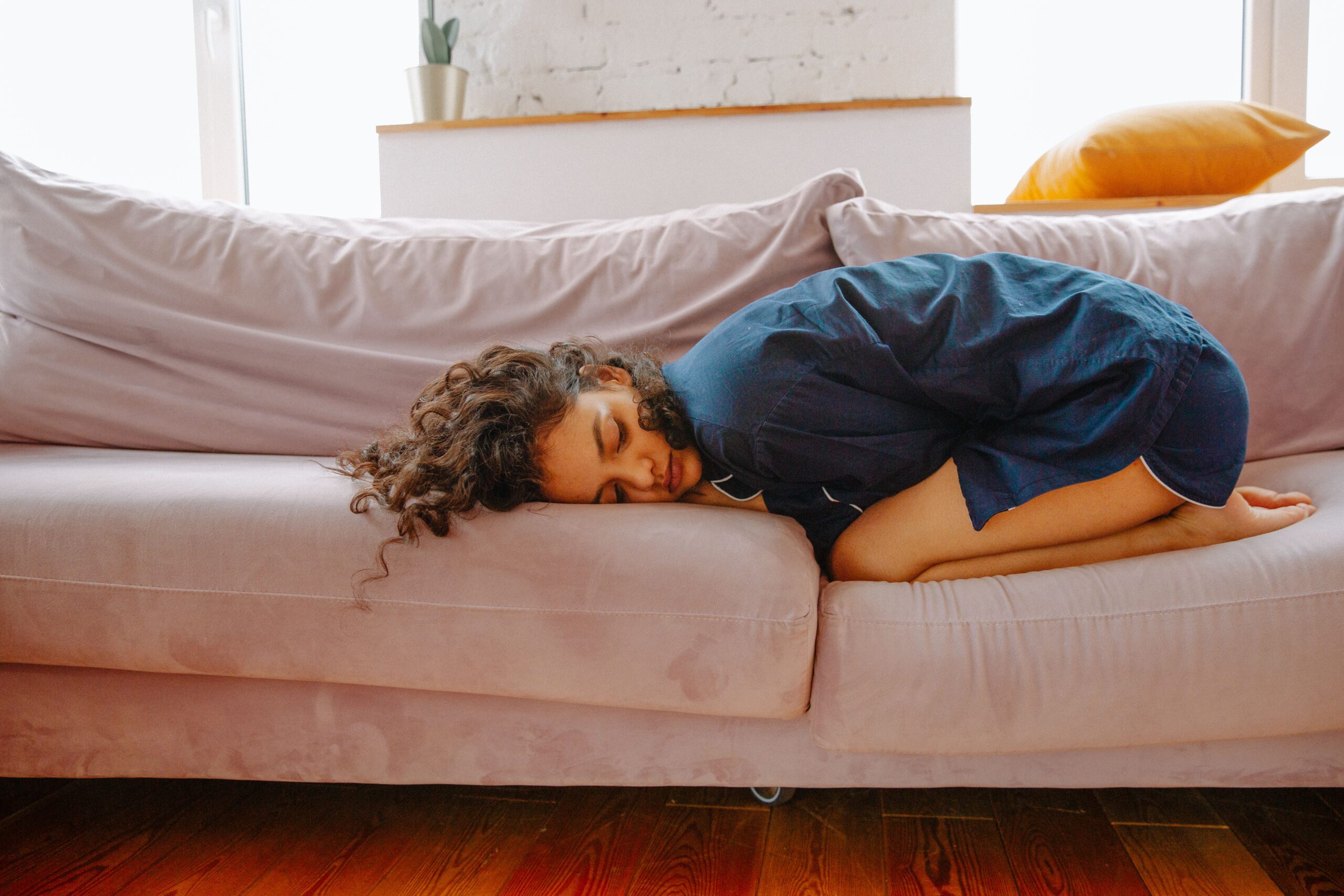 A woman with curly hair, showing signs of exhaustion, sleeps curled up on a light pink sofa in a bright room, wearing a blue shirt. A yellow cushion and a potted plant are visible in the background. Signalling the Link Between Trauma and Chronic Fatigue