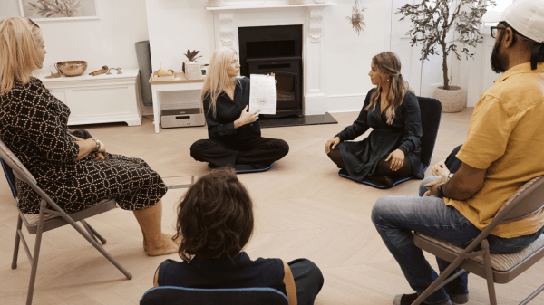 A group of five people sits in a circle indoors at the Trauma Clinic. One person in the centre holds up a piece of paper and speaks, while the others listen attentively. The room is decorated in a modern, minimalist style.
