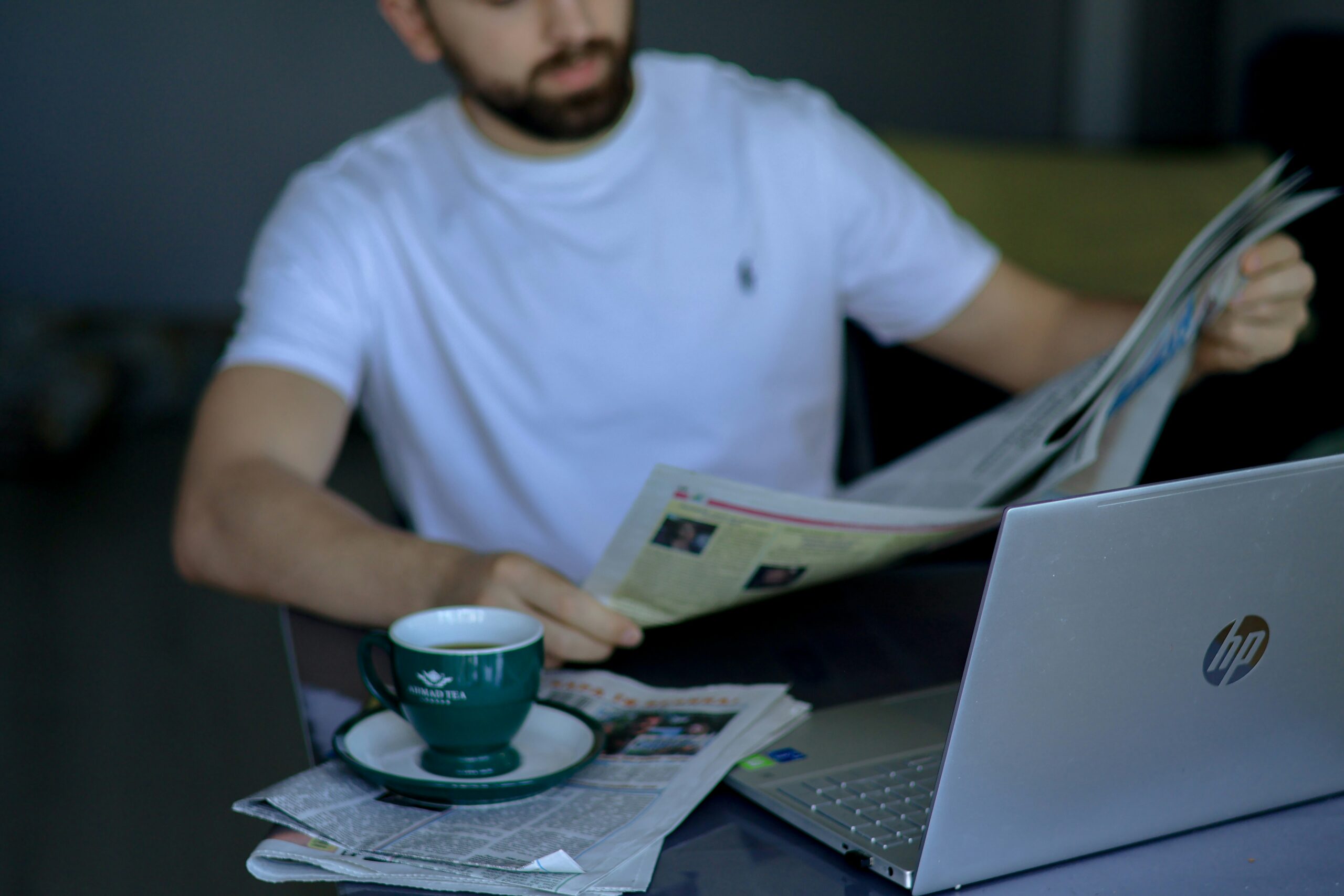 A person in a white shirt reads a newspaper at a table with an HP laptop, newspapers, and a green cup and saucer. Immersed in the latest global news, they sit thoughtfully whilst the background remains blurred.