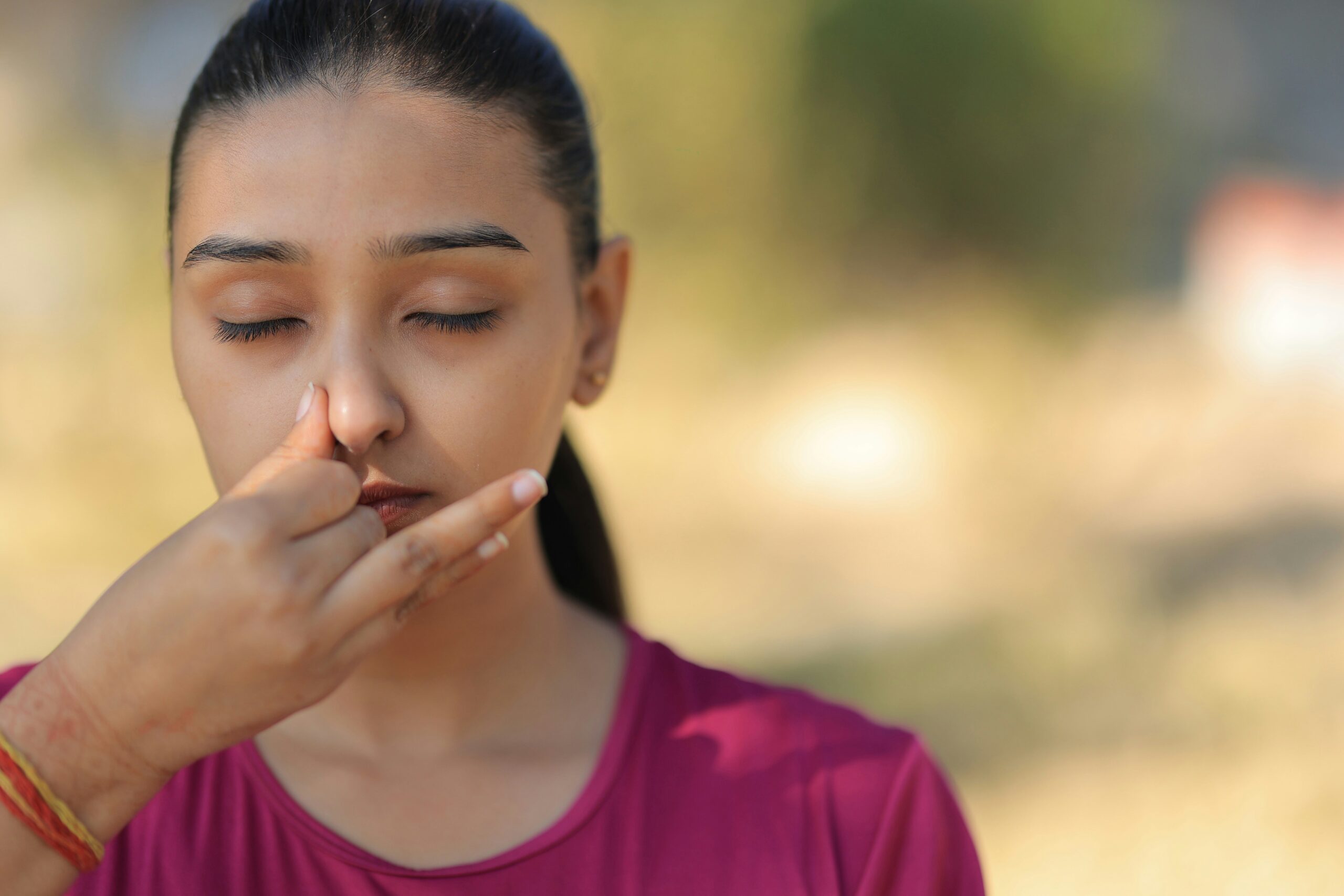 A woman with closed eyes practises breathwork and alternate nostril breathing outdoors, using her right hand to close one nostril. She wears a pink top and stands against a blurred background, focusing on the process of healing trauma.