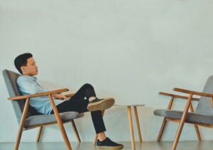 A man in a light blue shirt sits sideways in a wooden armchair with his legs crossed and one foot resting on another chair. There is a small round table between the two chairs, against a plain white wall, creating a quiet scene that subtly reminds us that helping others does not make us immune to trauma.