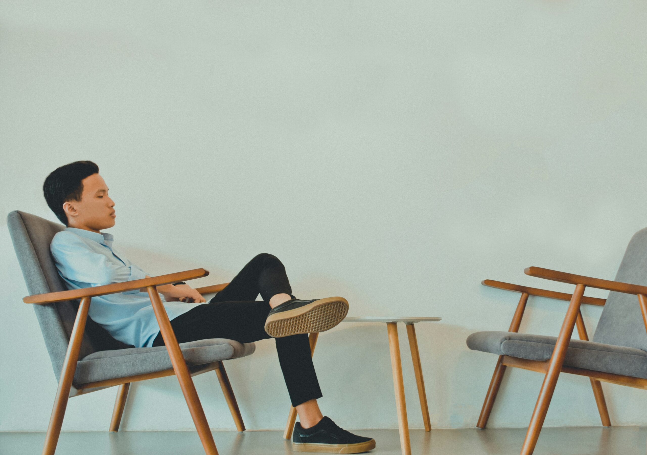 A man in a light blue shirt sits sideways in a wooden armchair with his legs crossed and one foot resting on another chair. There is a small round table between the two chairs, against a plain white wall, creating a quiet scene that subtly reminds us that helping others does not make us immune to trauma.