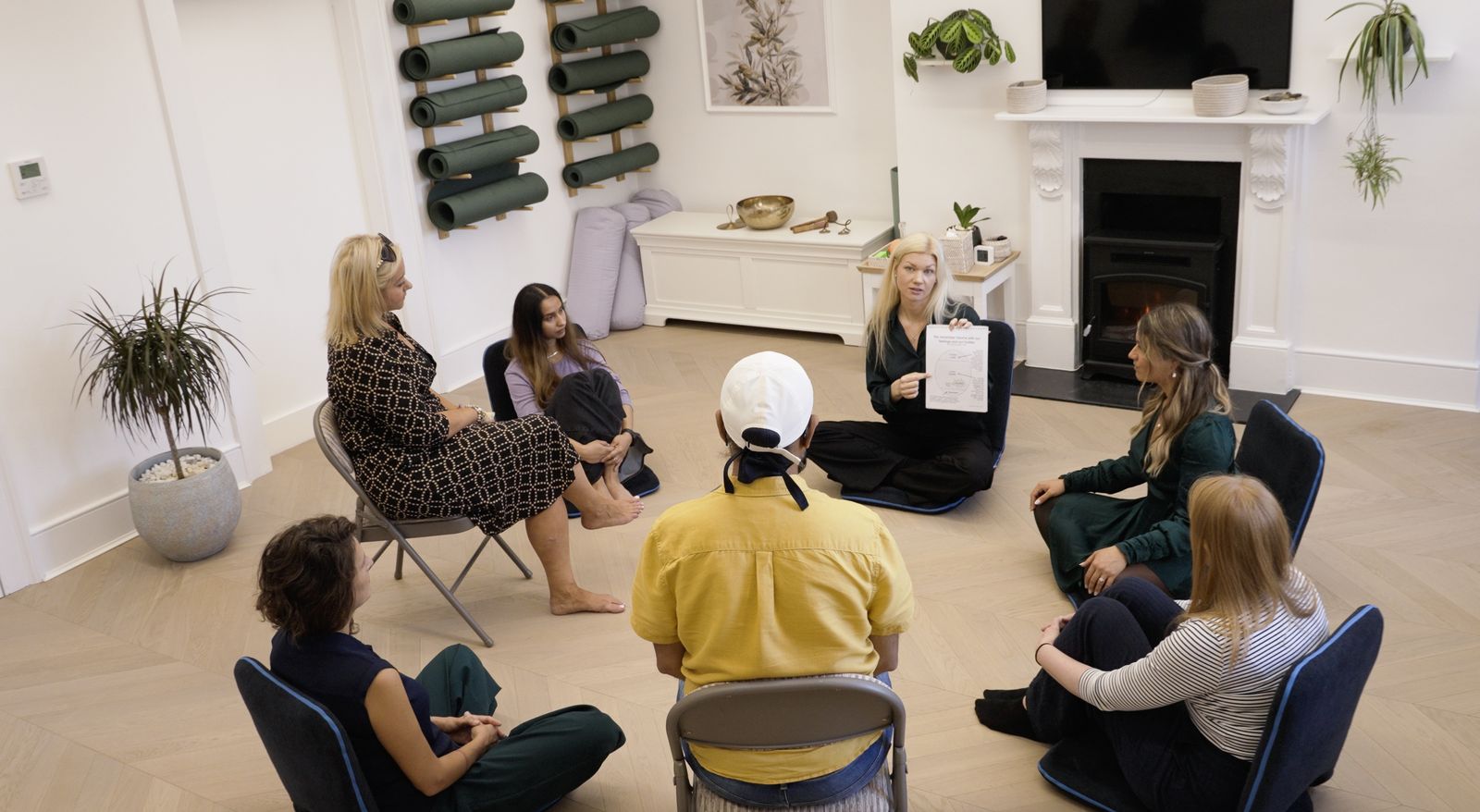 A group of seven people sit in a circle in a modern room at a mental health clinic; one woman holds up a document and speaks whilst others listen. Yoga mats are stacked on the wall, and plants decorate the space.
