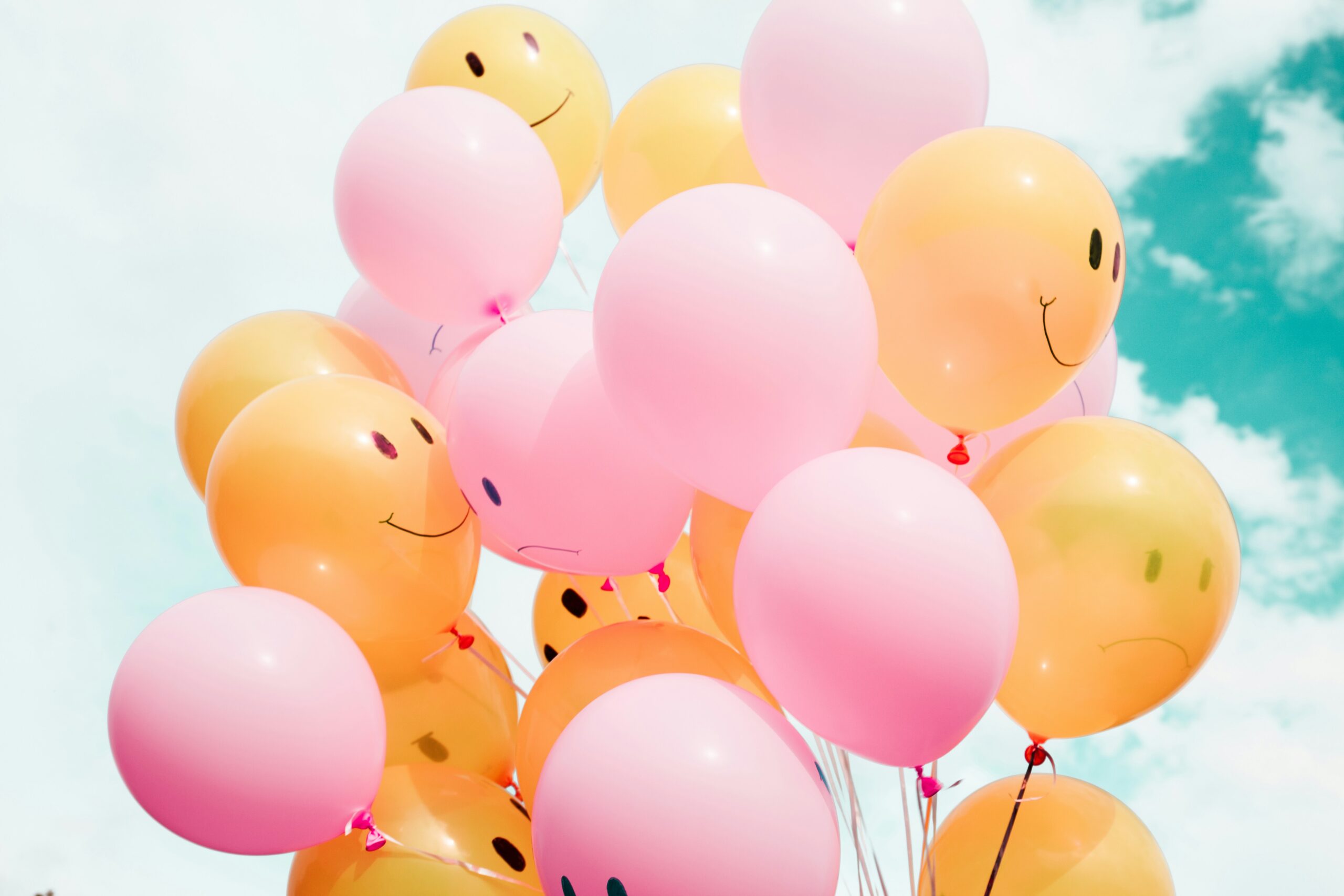 A bunch of yellow and pink balloons with smiley and neutral faces drawn on them float against a bright blue sky with scattered clouds, capturing the fleeting feelings of happiness and uncertainty that ripple through the nervous system as we quietly wonder, “Am I safe?”.