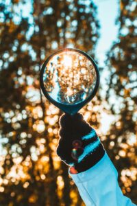 A hand in a black glove holds a magnifying glass up to sunlight, focusing on blurred trees in the background. The glass captures and intensifies the sun’s rays, creating a bright, glowing effect amid the foliage—as if lived reality is telling you there’s more to see beyond what first meets the eye.