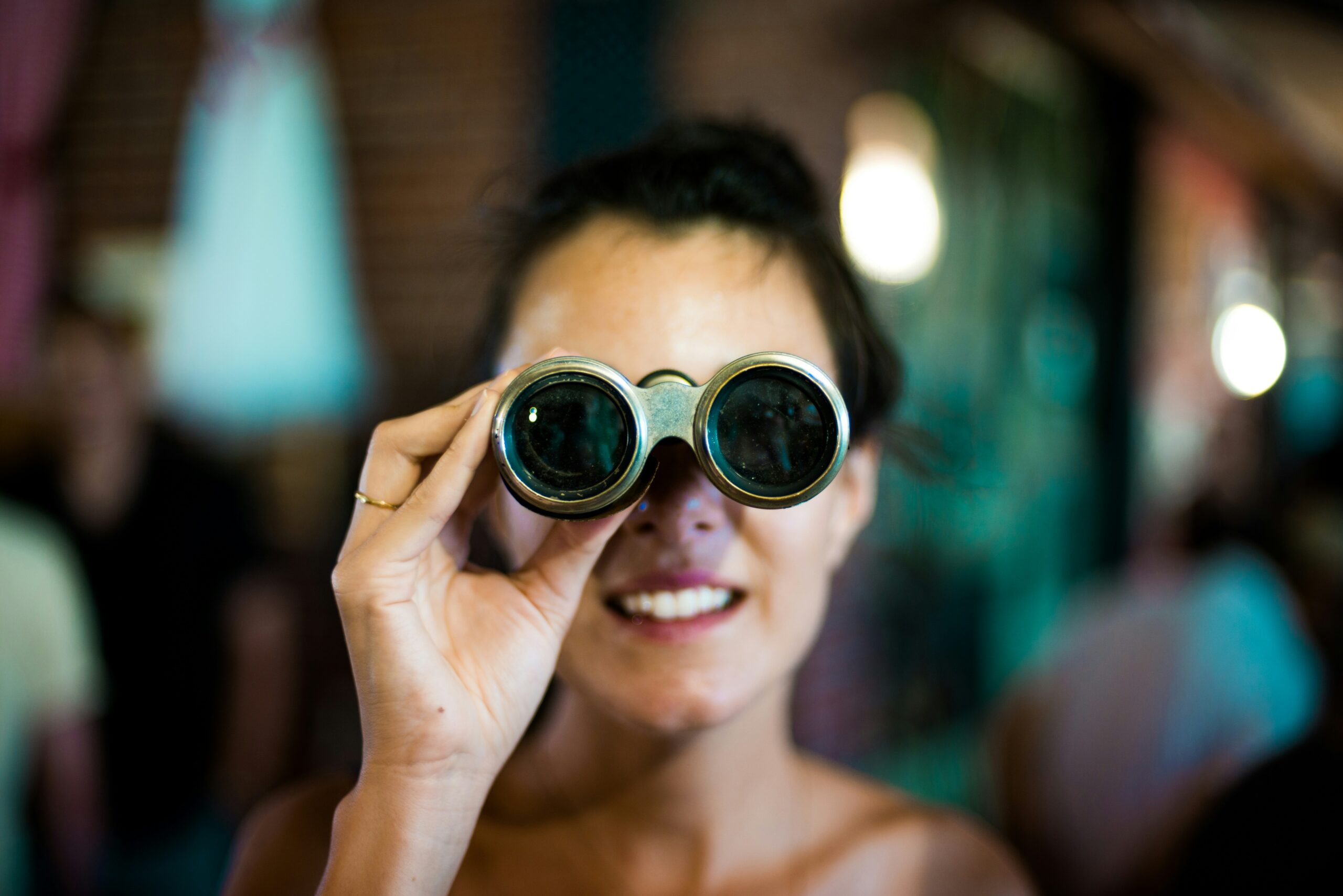A person smiles whilst holding binoculars up to their eyes, looking directly at the camera as if engaged in trigger identification. The background is blurred, focussing attention on the face and binoculars.