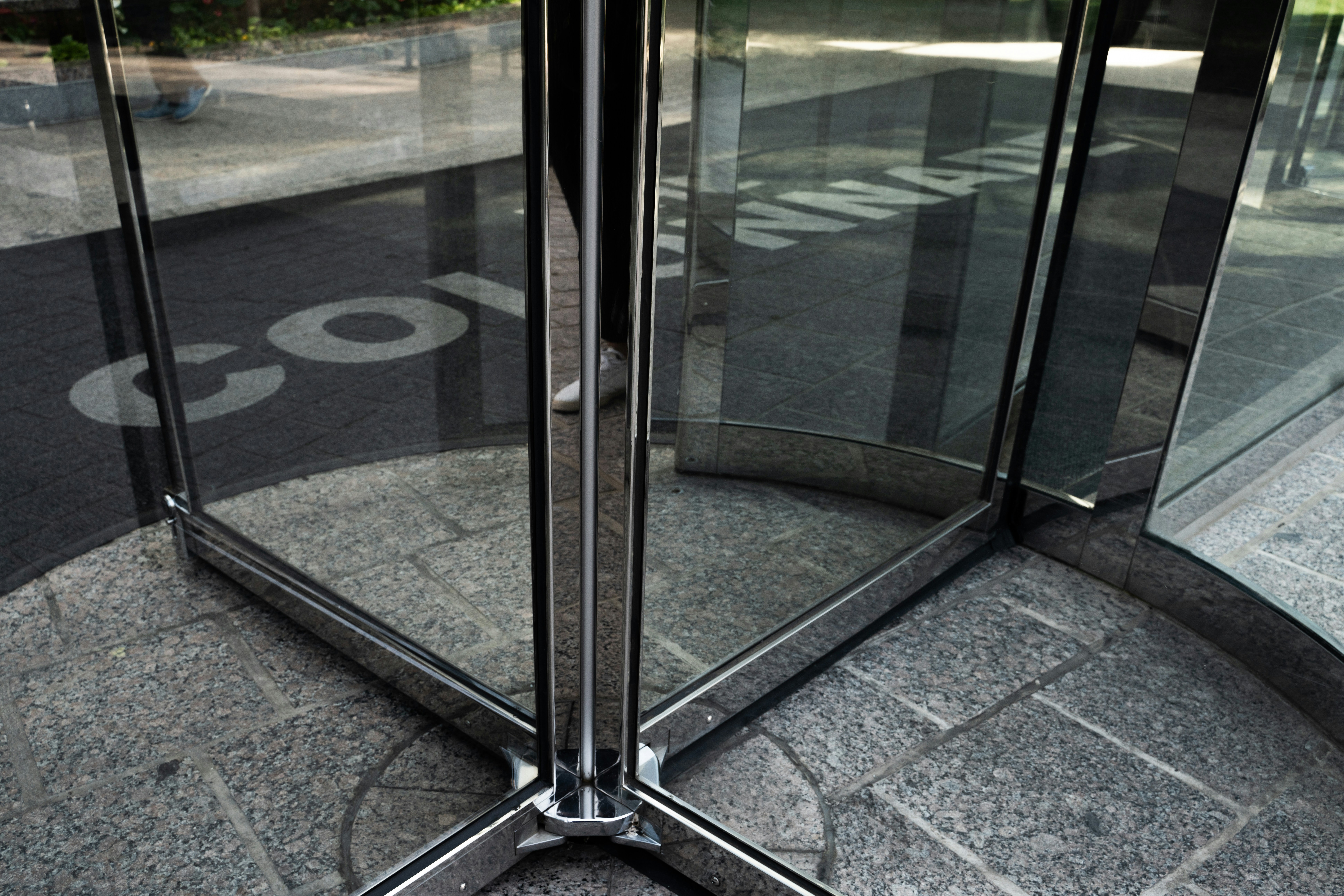 Glass revolving door viewed from inside, showing reflections and stone floor tiles—a subtle metaphor for the revolving door of treatment often experienced during mental health relapse—with partial white text visible on the dark doormat outside.