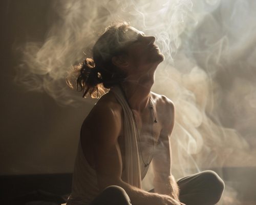 A person with long hair sits cross-legged in a dimly lit room in Oxfordshire, surrounded by swirling smoke, tilting their head back with eyes closed, appearing calm and contemplative—perhaps reflecting on their trauma recovery journey.