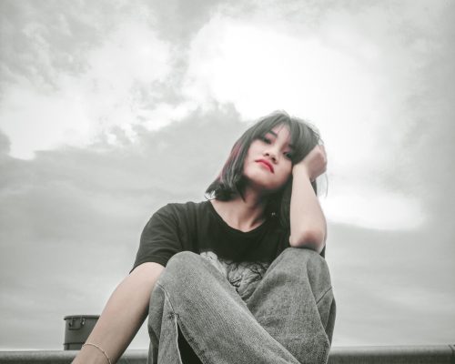 A woman with short dark hair sits outdoors in Oxfordshire, resting her head on her hand and looking contemplative. She wears a black t-shirt and jeans, reflecting on trauma recovery under a cloudy sky.